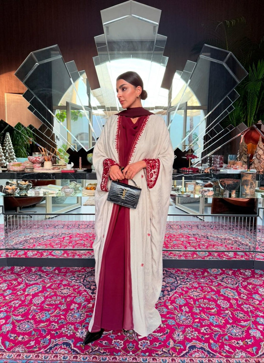 Woman wearing Harir Kubra Ivory Abaya with burgundy embroidery on sleeves and front edges in elegant indoor setting