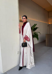 Woman wearing Harir Kubra Ivory Abaya with burgundy embroidery on sleeves and front edges in elegant indoor setting