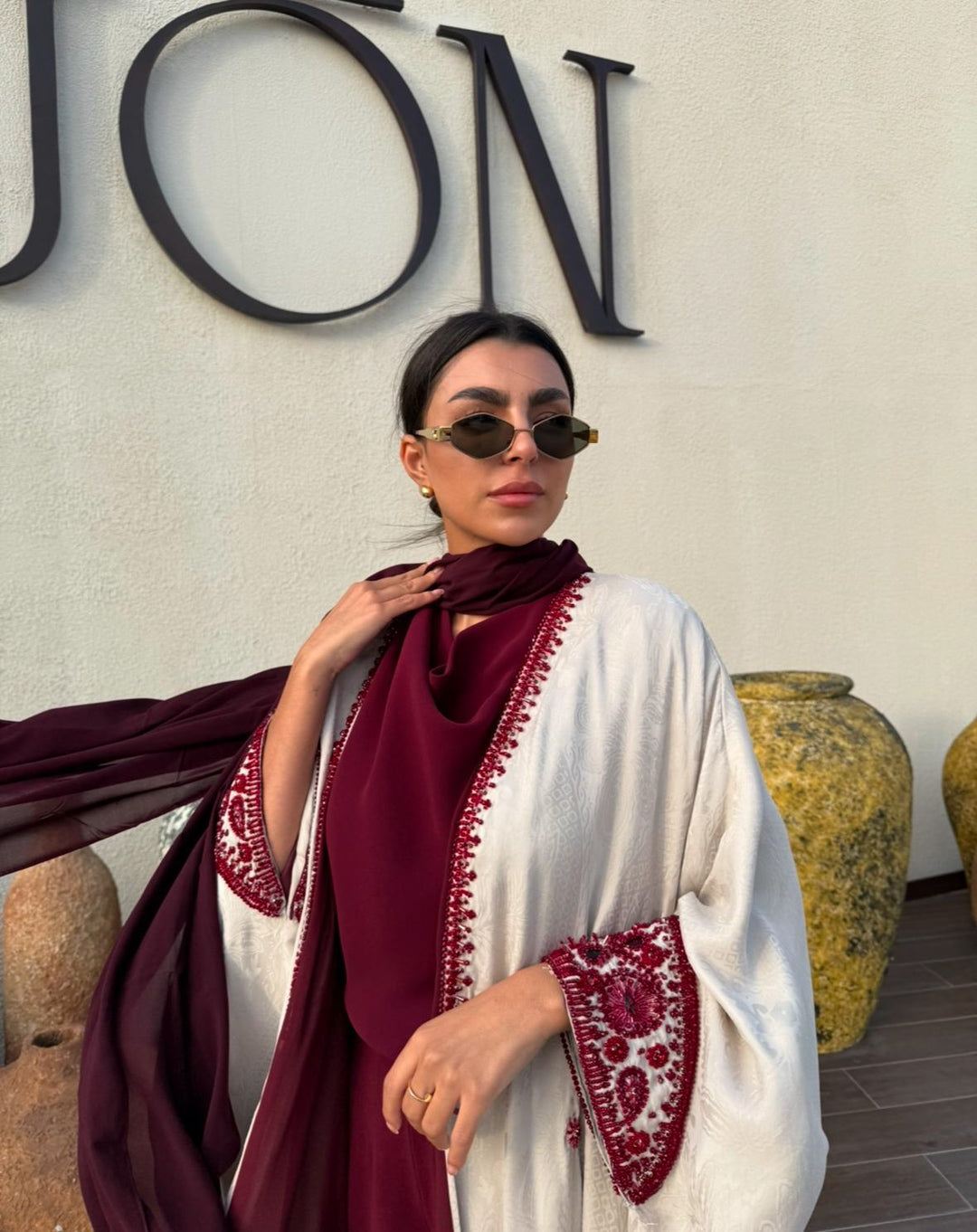 Woman wearing Harir Kubra Ivory Abaya with burgundy embroidery on sleeves and front edges in elegant indoor setting