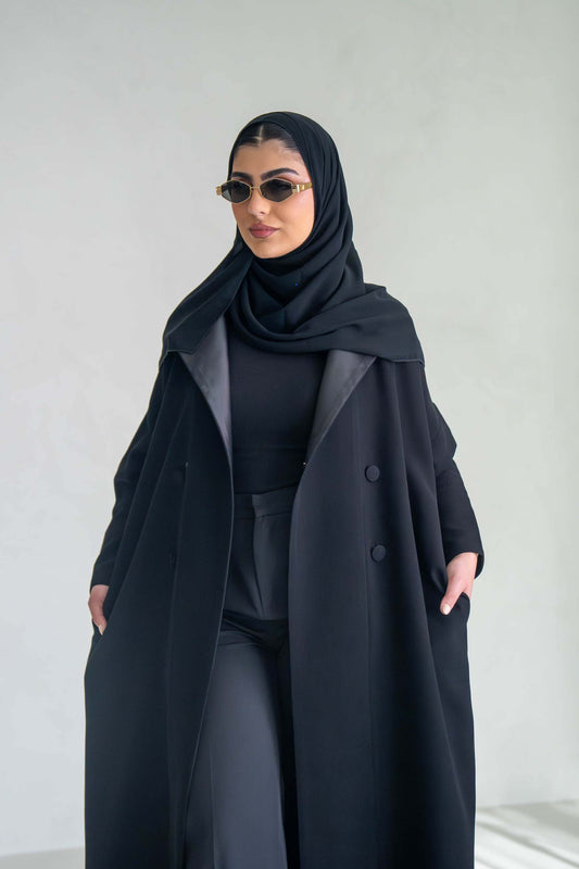 Woman wearing thick black crepe abaya with sunglasses and hijab against a plain background