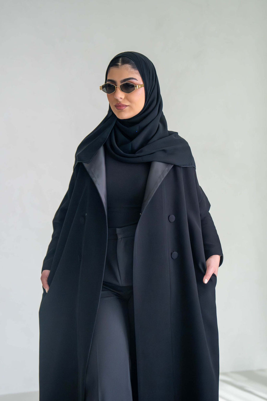Woman wearing thick black crepe abaya with sunglasses and hijab against a plain background