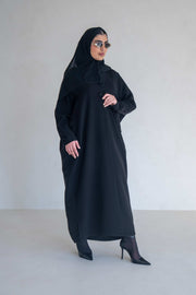 Woman wearing a classic black abaya with wide sleeves and side pockets, styled with black heels and sunglasses.