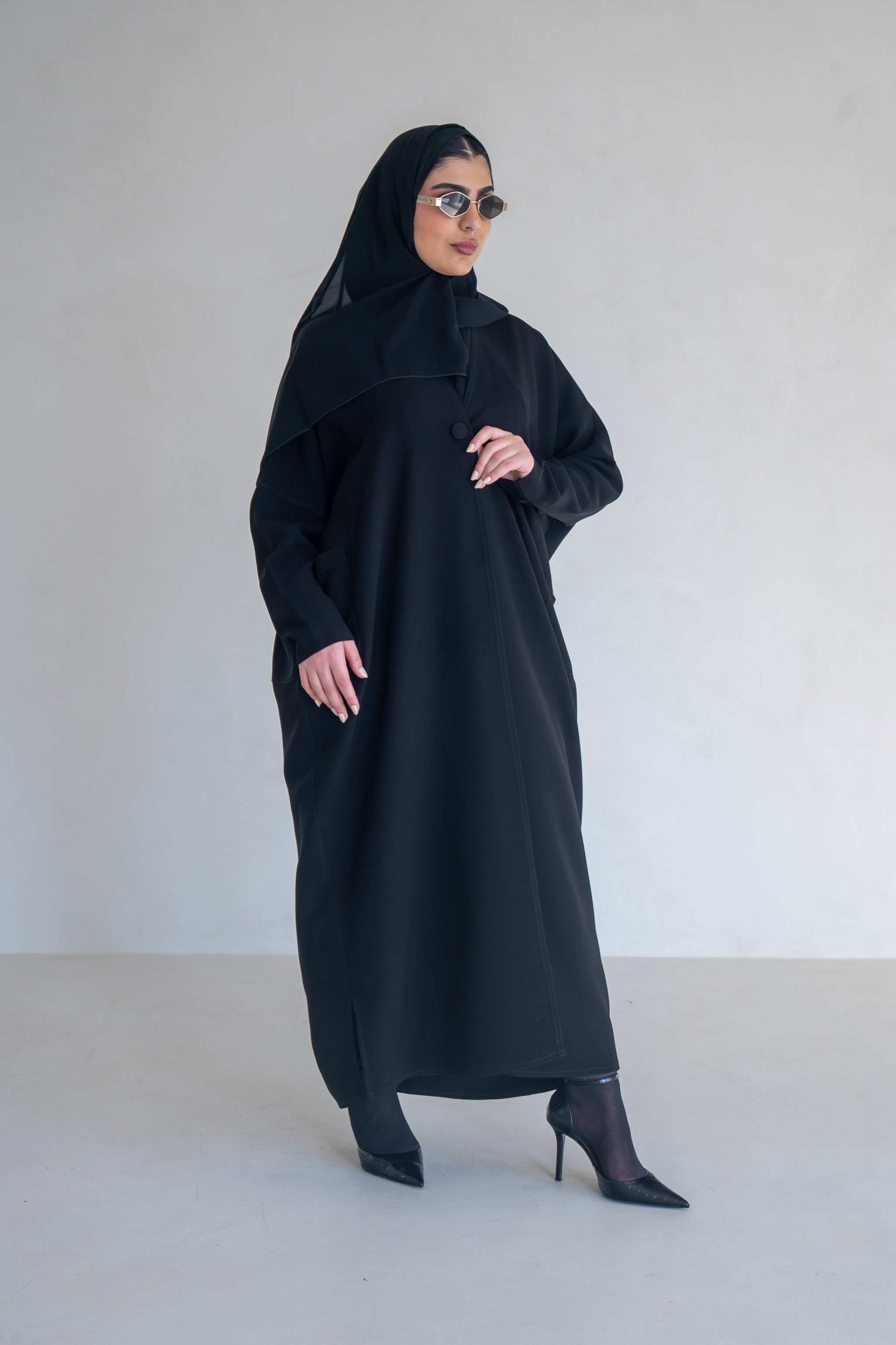 Woman wearing a classic black abaya with wide sleeves and side pockets, styled with black heels and sunglasses.
