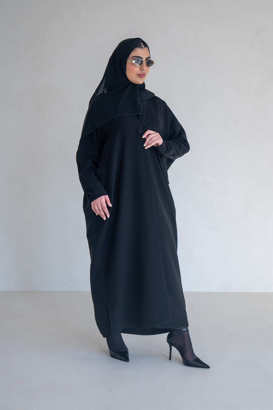 Woman wearing a classic black abaya with wide sleeves and side pockets, styled with black heels and sunglasses.
