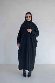 woman wearing classic black abaya with wide sleeves and side pockets standing against plain background