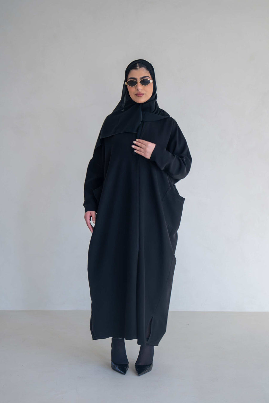 woman wearing classic black abaya with wide sleeves and side pockets standing against plain background