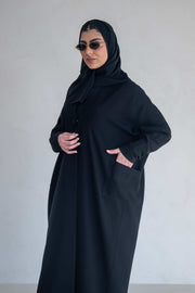woman wearing classic black abaya with wide sleeves and side pockets standing against plain background