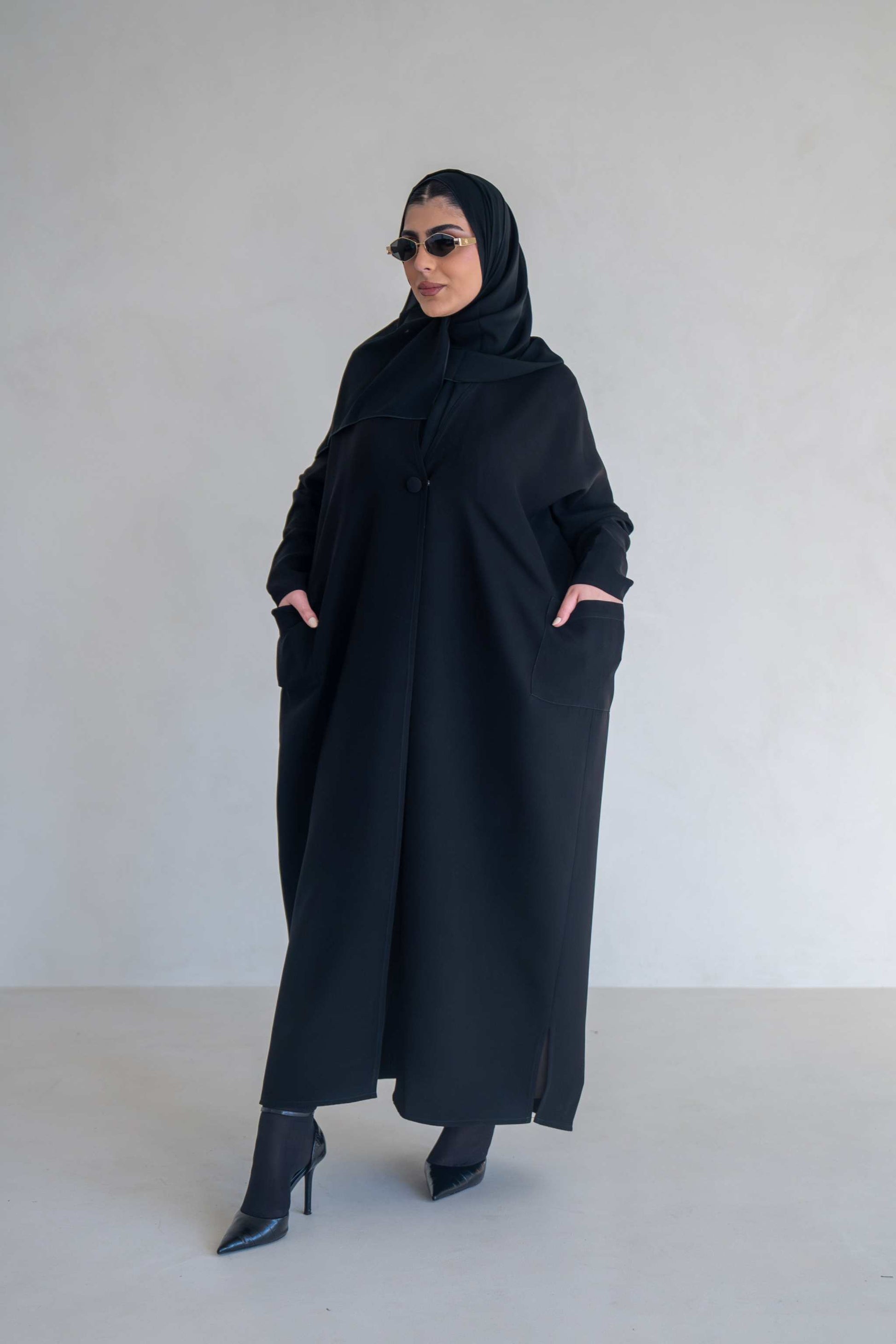 woman wearing classic black abaya with wide sleeves and side pockets standing against plain background