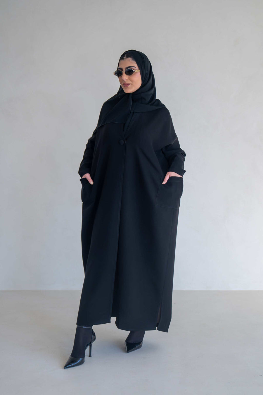 woman wearing classic black abaya with wide sleeves and side pockets standing against plain background