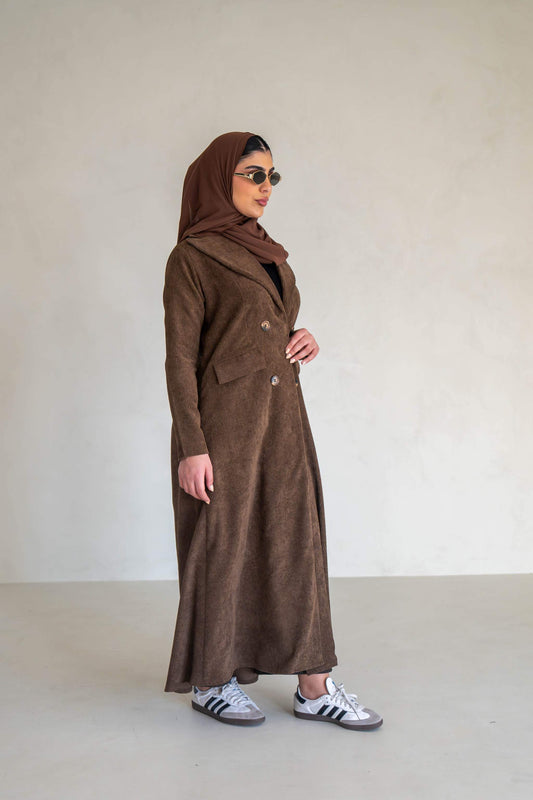 Woman wearing a brown velvet corduroy abaya with buttons and false pockets standing against a plain background