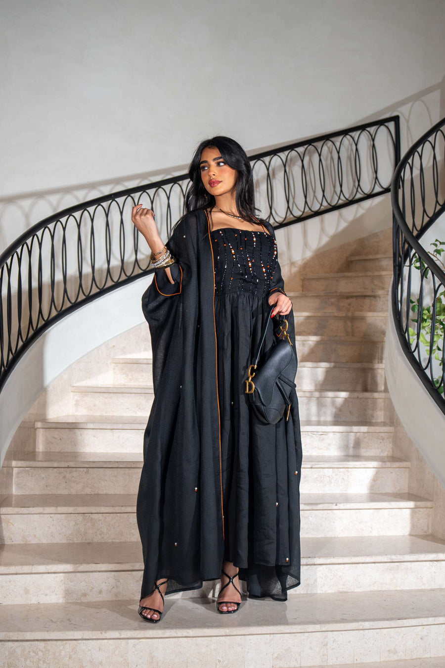 Woman wearing a black gathered dress with matching bisht featuring bold orange piping and scattered embellishments on stairs