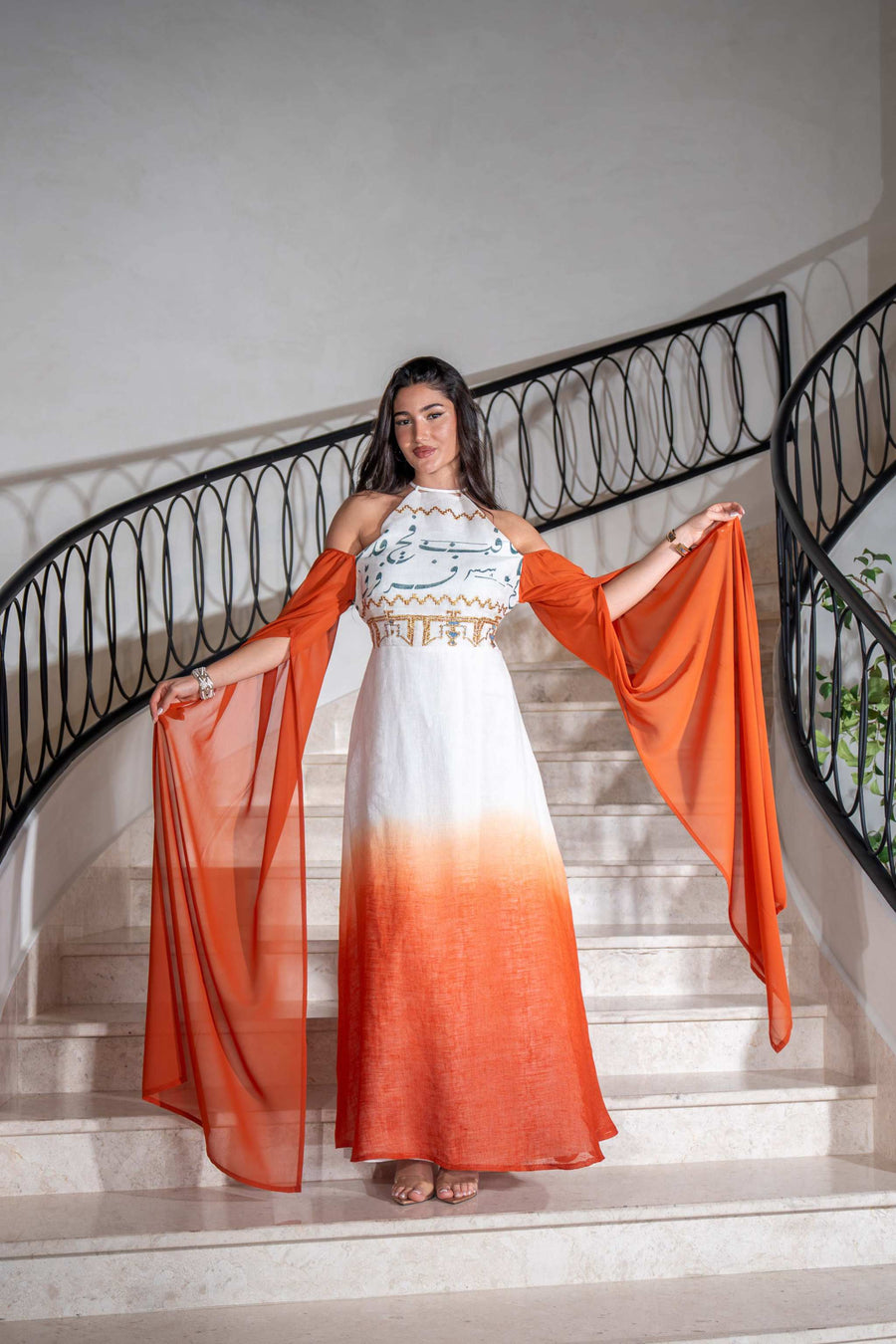 Woman wearing a Tangerine Ombre cold-shoulder dress with dramatic cape sleeves and hand embroidery on the bodice.