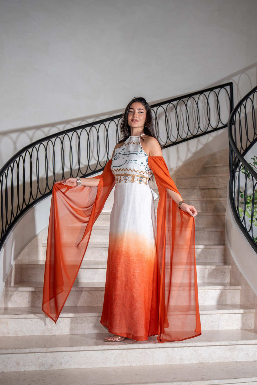 Woman wearing a Tangerine Ombre cold-shoulder dress with dramatic cape sleeves and hand embroidery, standing on staircase.
