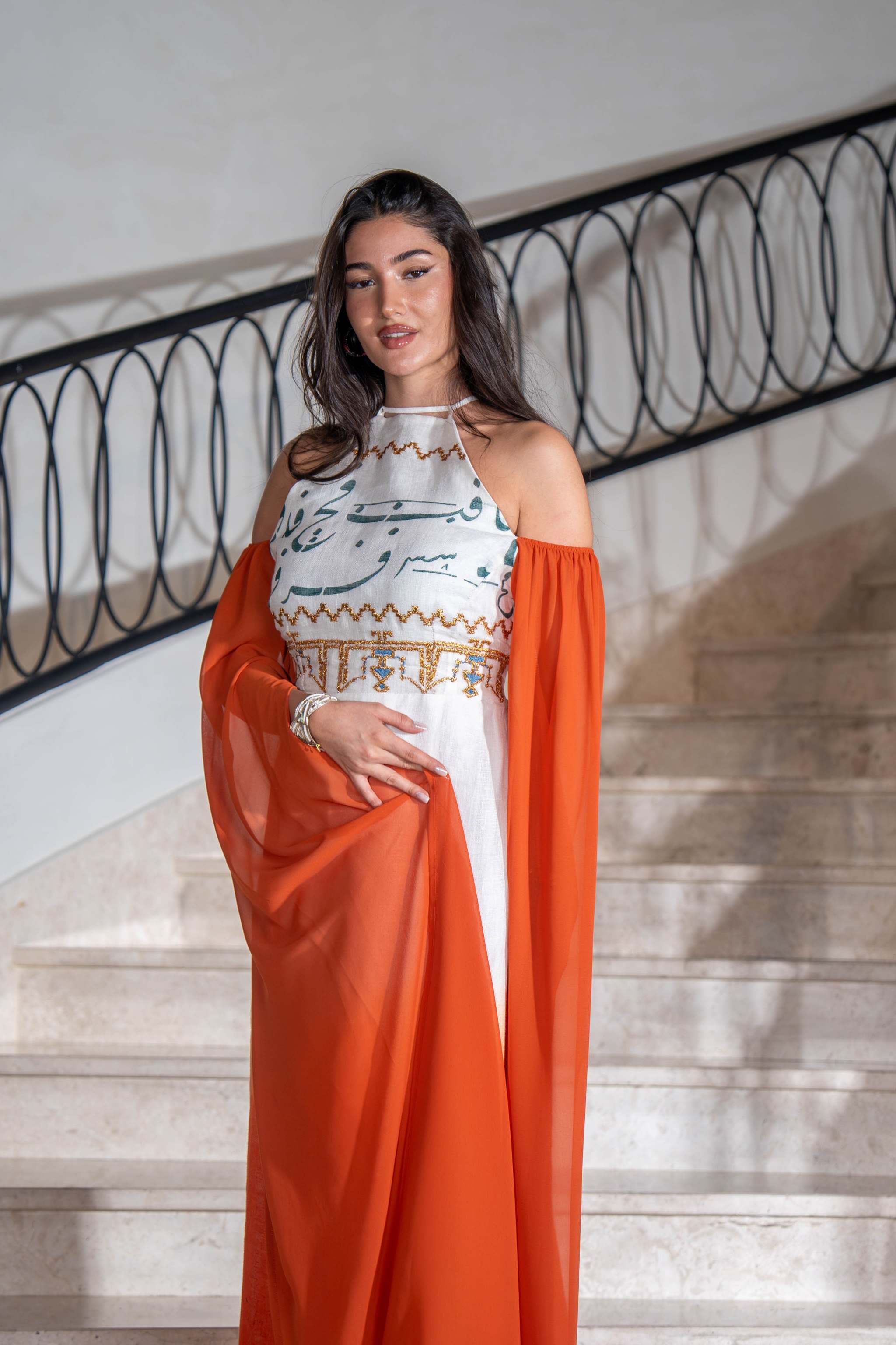Woman wearing a tangerine and white ombre cold-shoulder dress with dramatic cape sleeves and hand-embroidered bodice standing on stairs.