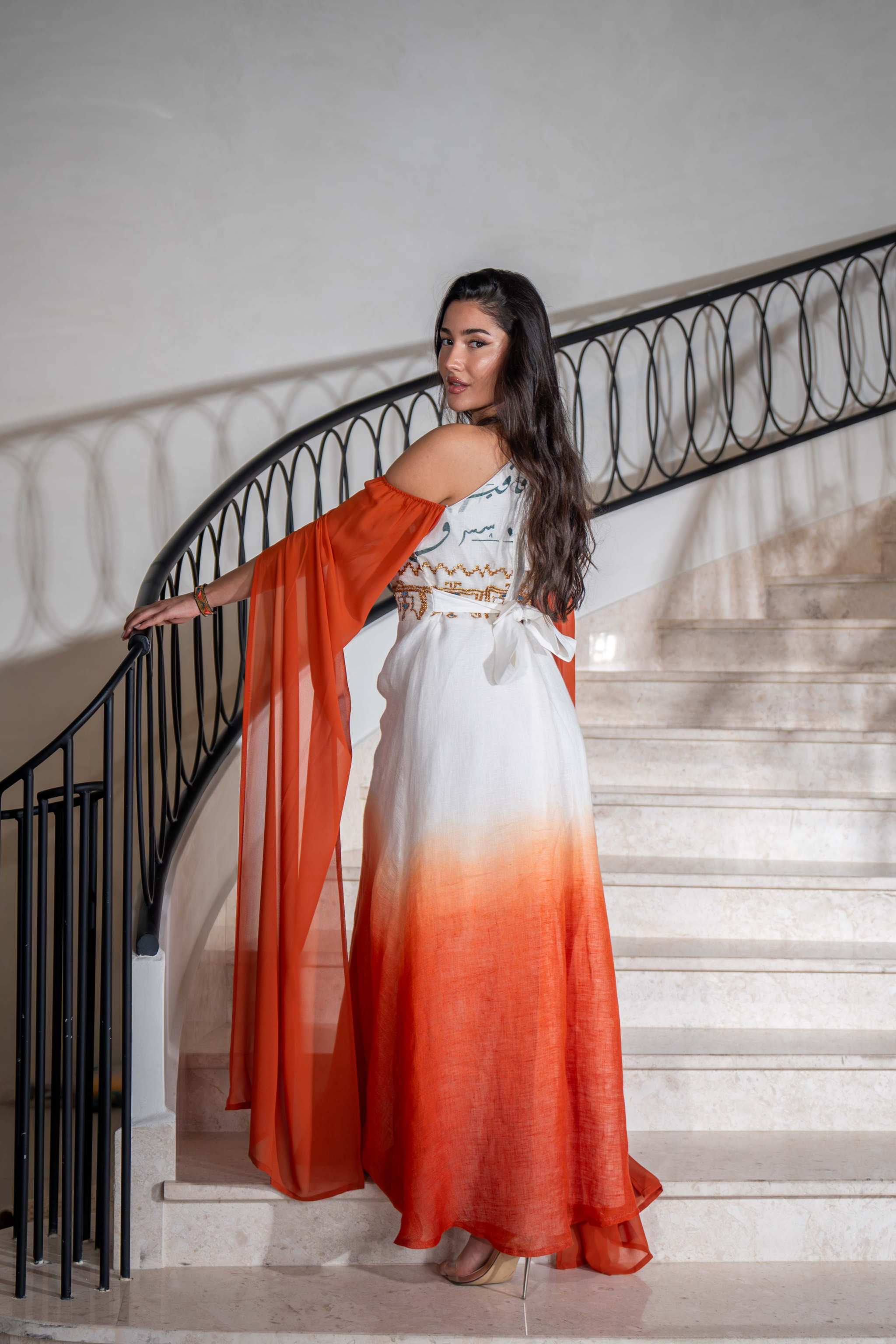 Woman wearing a Tangerine Ombre cold-shoulder dress with flowing cape sleeves and embroidered bodice standing on stairs