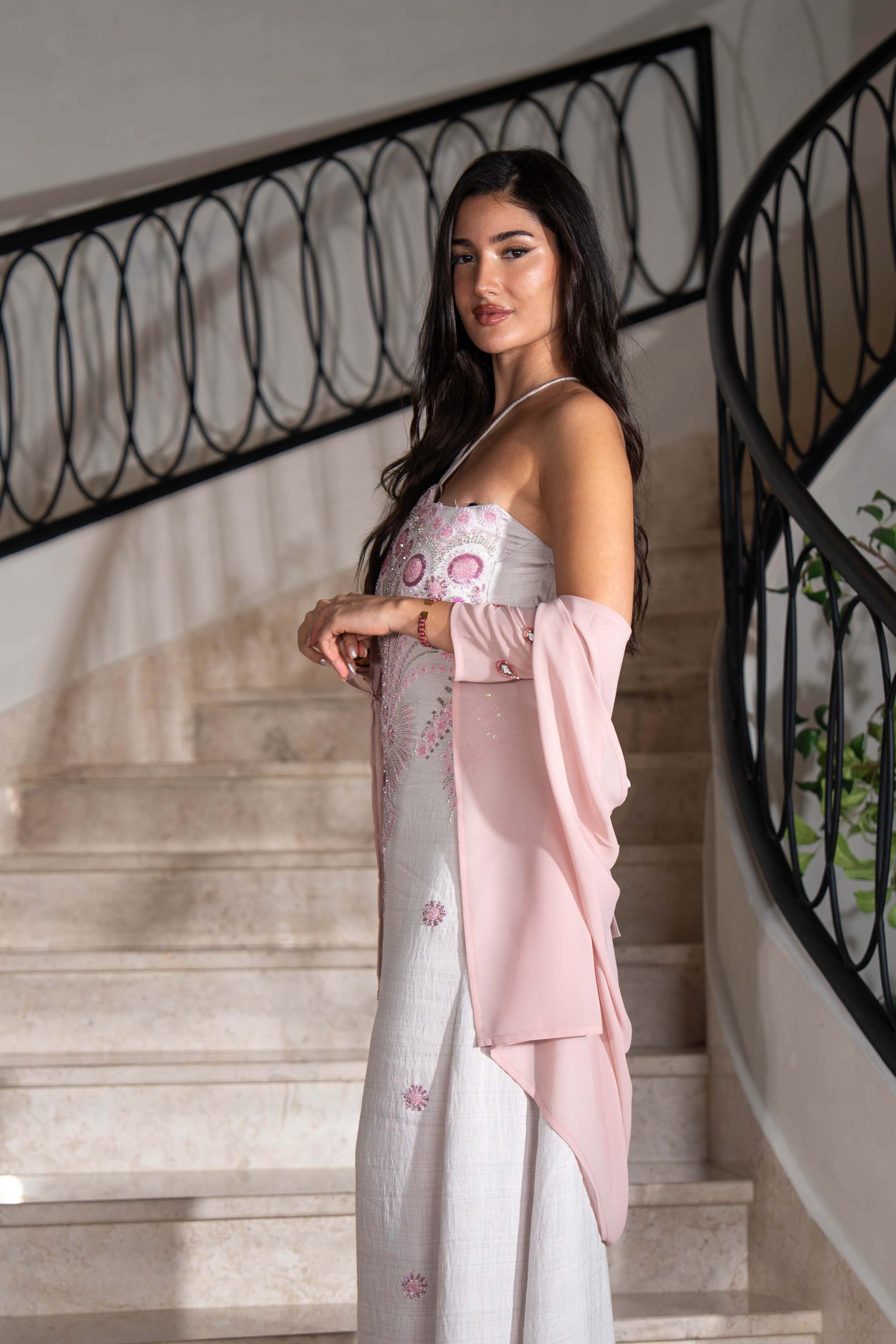 Elegant white one-piece dress with pink embroidery and blush pink cape sleeves worn by a woman on stairs