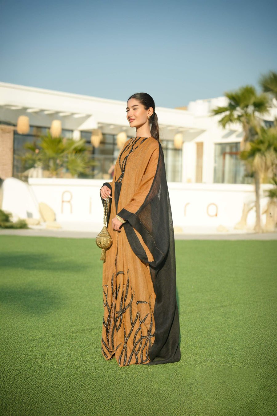 Woman wearing a black sleeveless jumpsuit layered with a golden-brown embroidered linen bisht holding a decorative gold clutch outdoors.