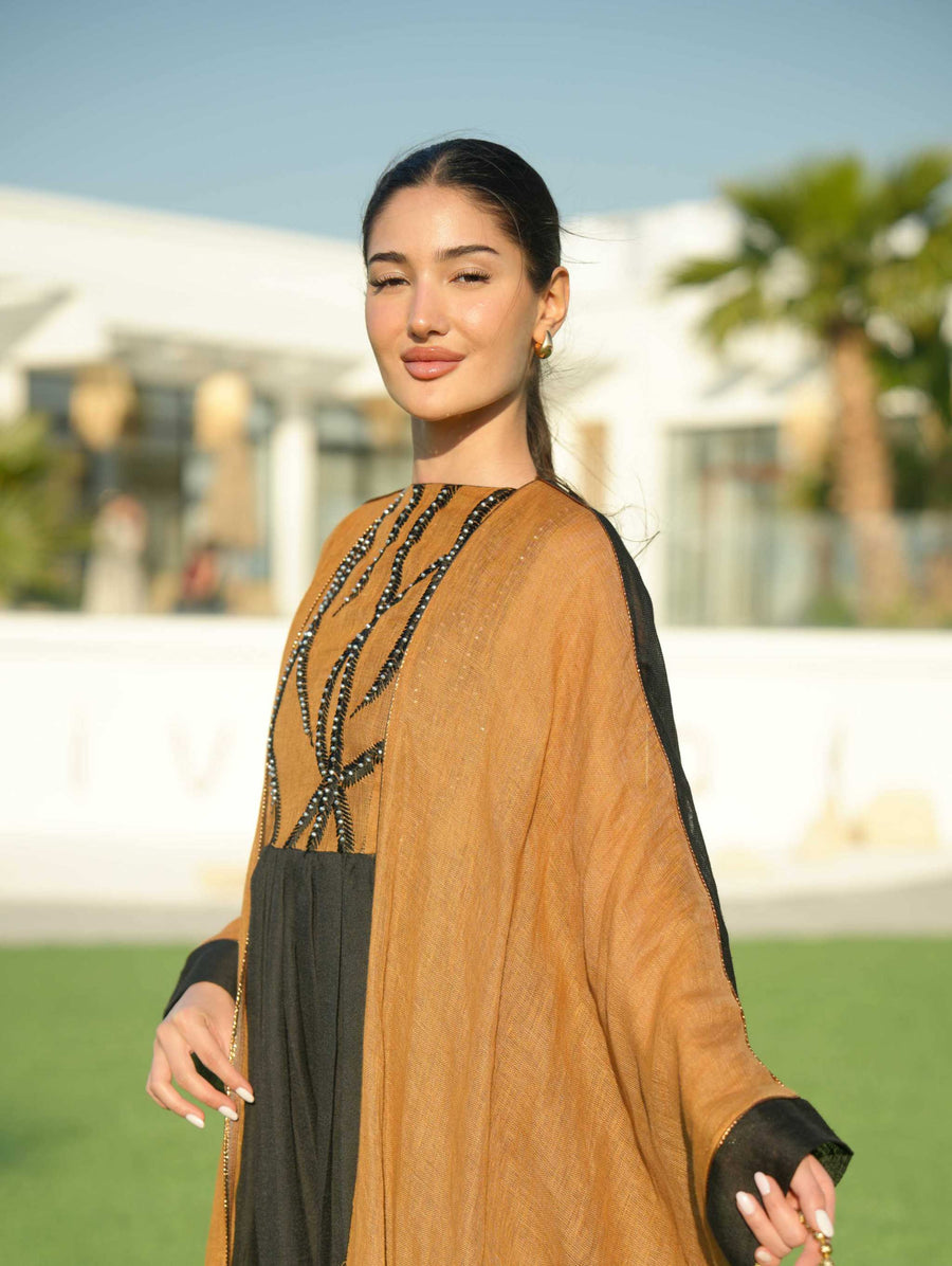 Woman wearing a black sleeveless jumpsuit layered with a golden-brown embroidered linen bisht holding a decorative gold clutch outdoors.