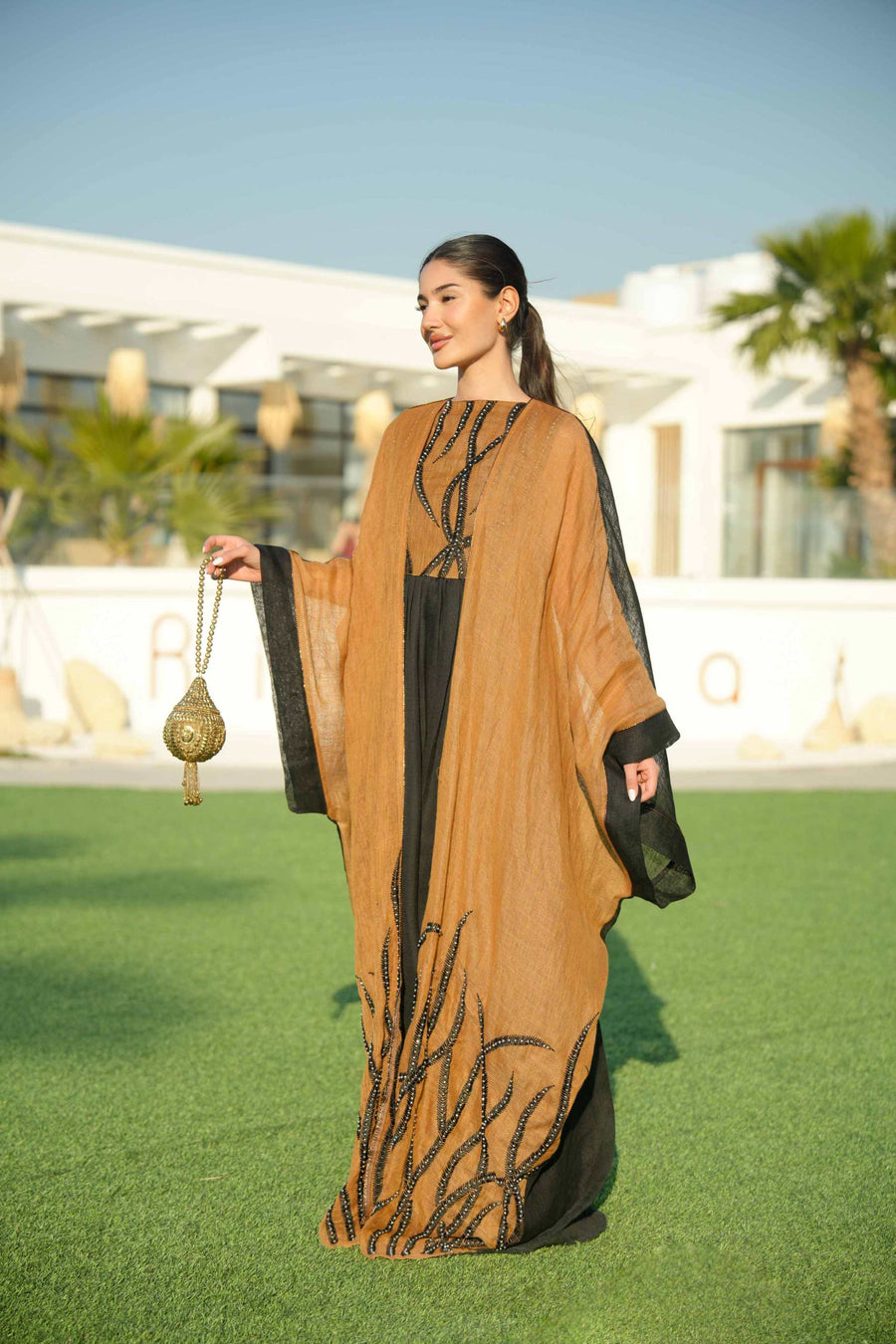 Woman wearing a black sleeveless jumpsuit layered with a golden-brown embroidered linen bisht holding a decorative gold clutch outdoors.