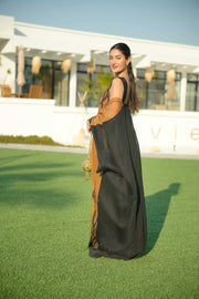 Woman wearing a black sleeveless jumpsuit layered with a golden-brown embroidered linen bisht holding a decorative gold clutch outdoors.