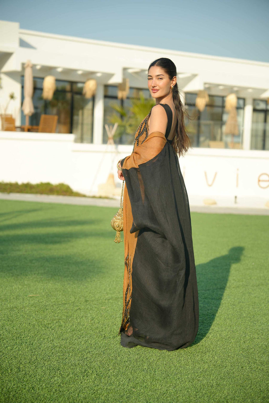 Woman wearing a black sleeveless jumpsuit layered with a golden-brown embroidered linen bisht holding a decorative gold clutch outdoors.