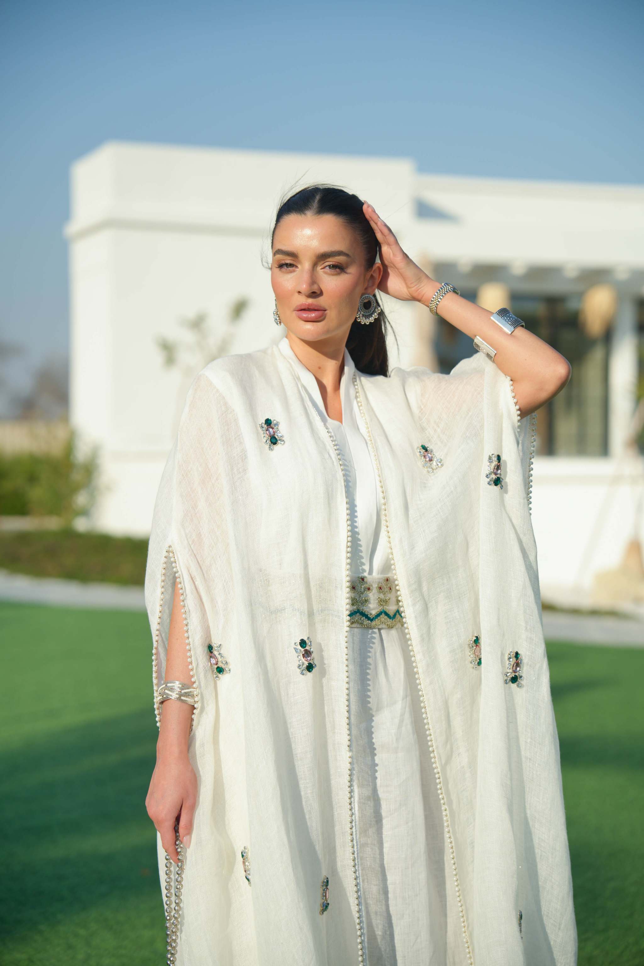 Woman wearing an elegant all-white linen jumpsuit with a sheer embroidered bisht featuring green and black floral motifs and side slits, styled for daytime or festive occasions.