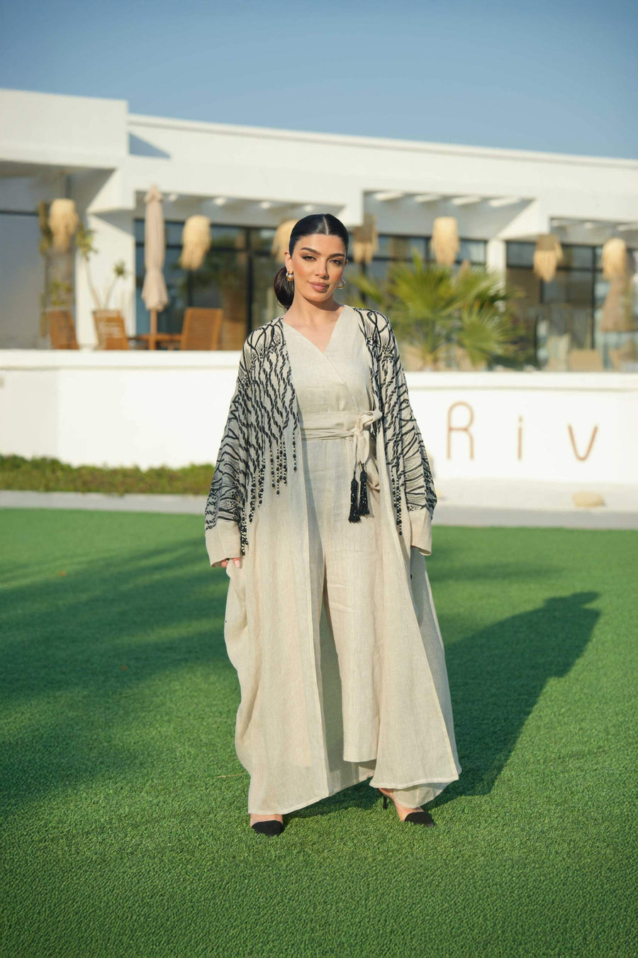 Woman wearing a beige linen jumpsuit with a waist tie and a black-embroidered bisht featuring cascading threadwork, standing outdoors on green grass.