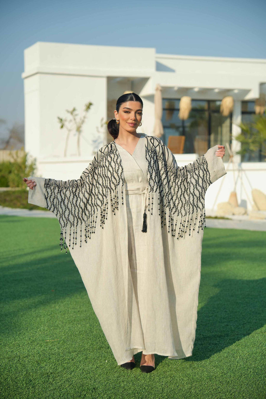 Woman wearing a beige linen jumpsuit with a waist tie and a black-embroidered bisht featuring cascading threadwork, standing outdoors on green grass.