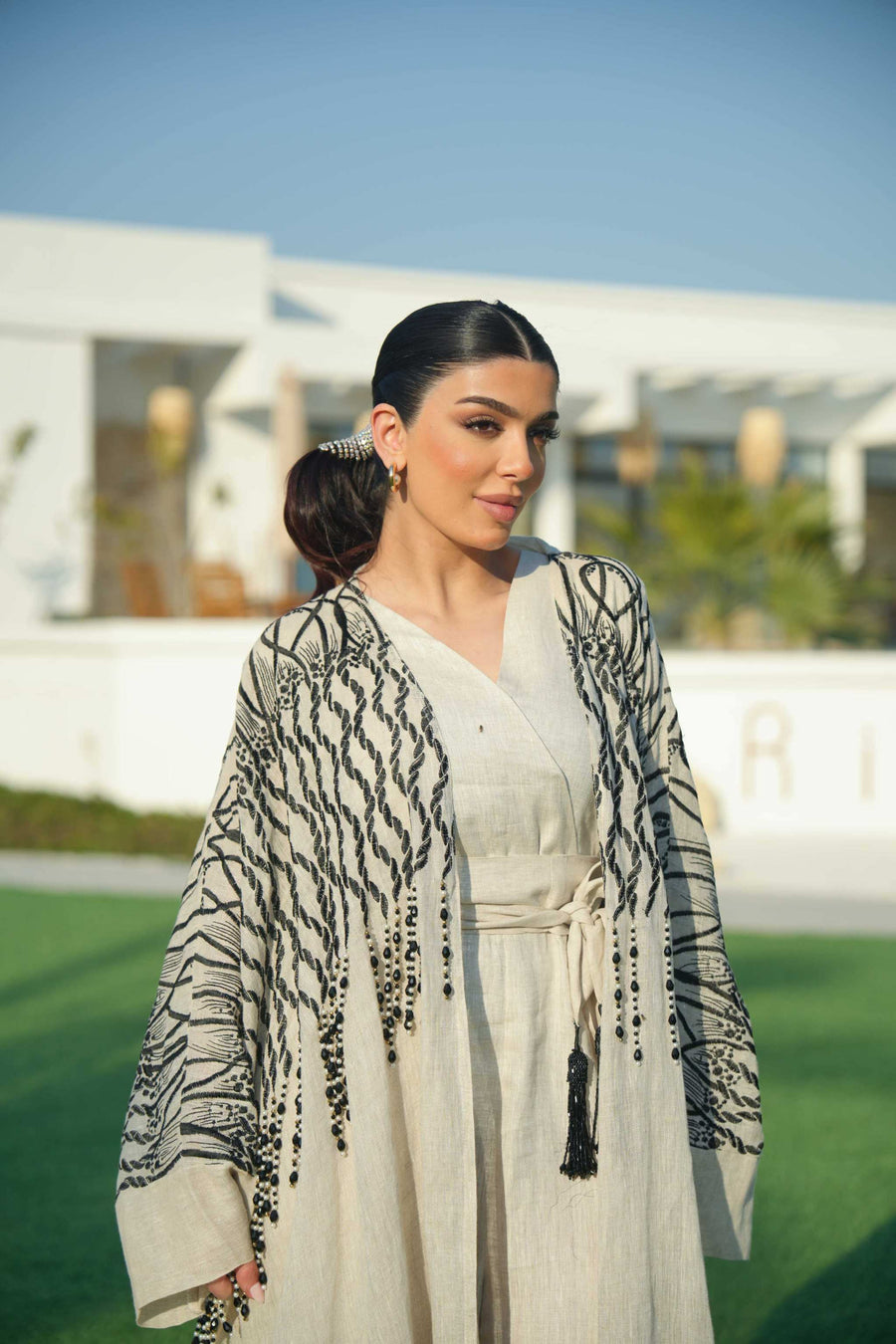 Woman wearing a beige linen jumpsuit with a waist tie and a black-embroidered bisht featuring cascading threadwork, standing outdoors on green grass.