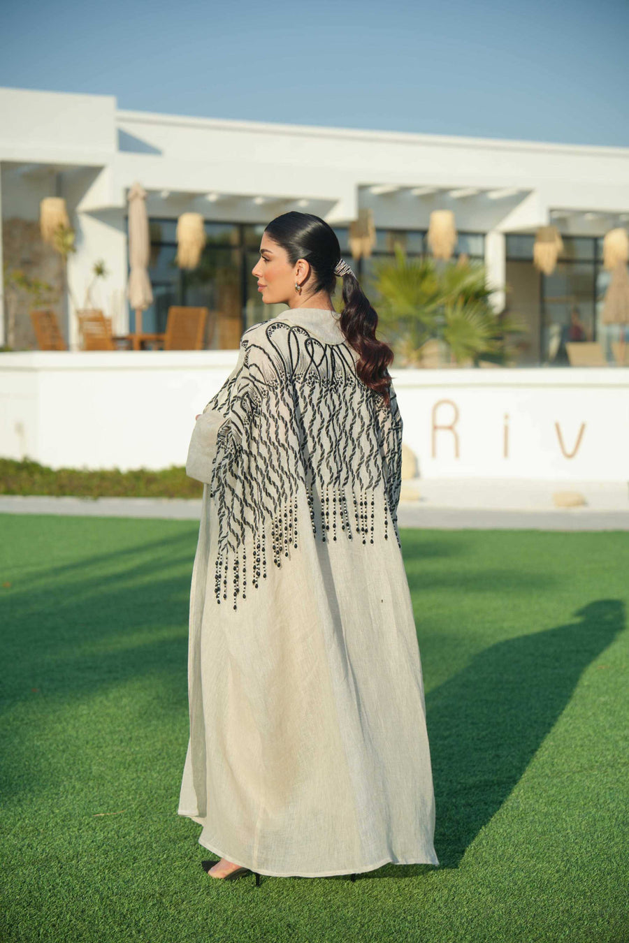 Woman wearing a beige linen jumpsuit with a waist tie and a black-embroidered bisht featuring cascading threadwork, standing outdoors on green grass.