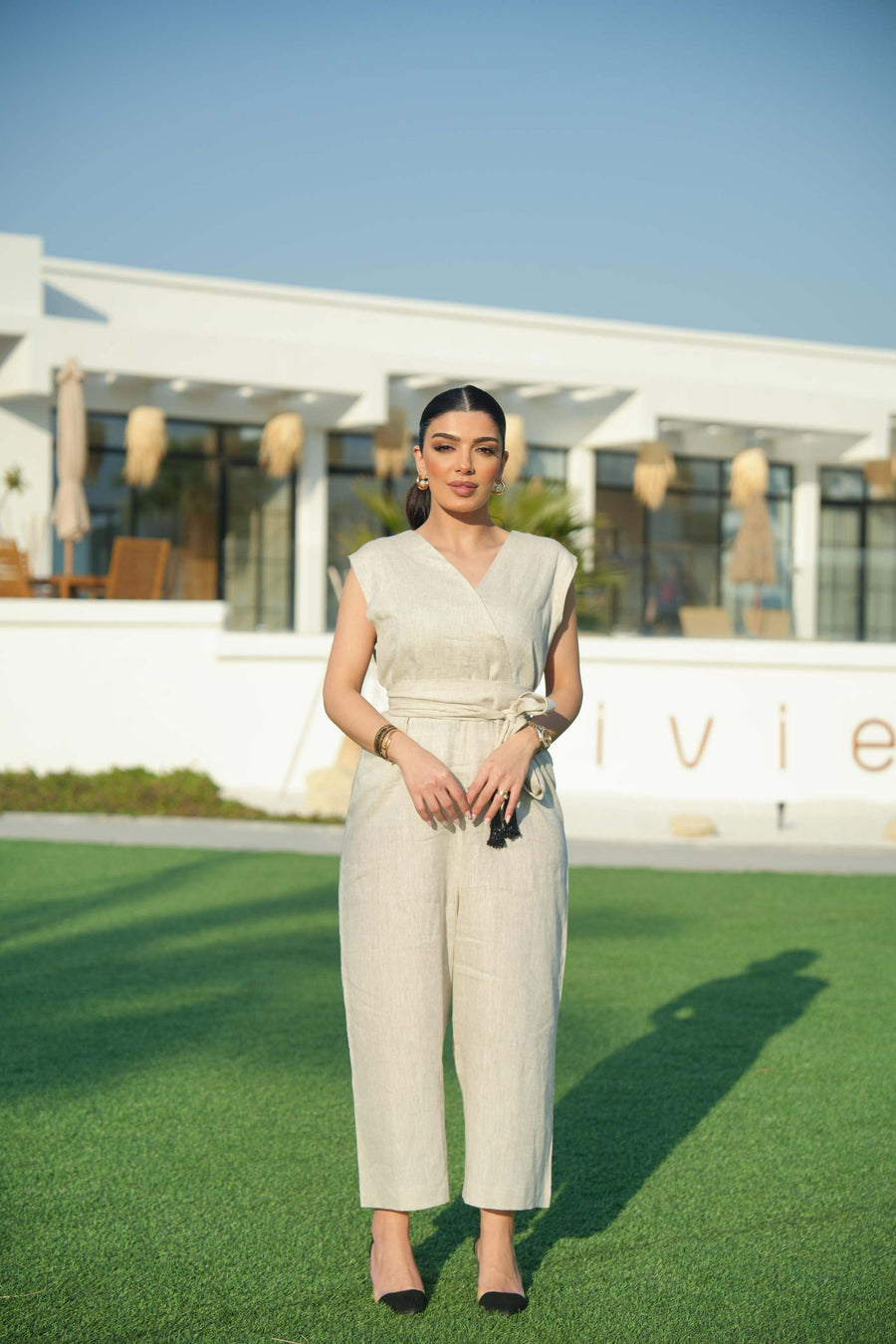 Woman wearing a beige linen jumpsuit with a waist tie and a black-embroidered bisht featuring cascading threadwork, standing outdoors on green grass.