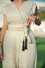 Woman wearing a beige linen jumpsuit with a waist tie and a black-embroidered bisht featuring cascading threadwork, standing outdoors on green grass.
