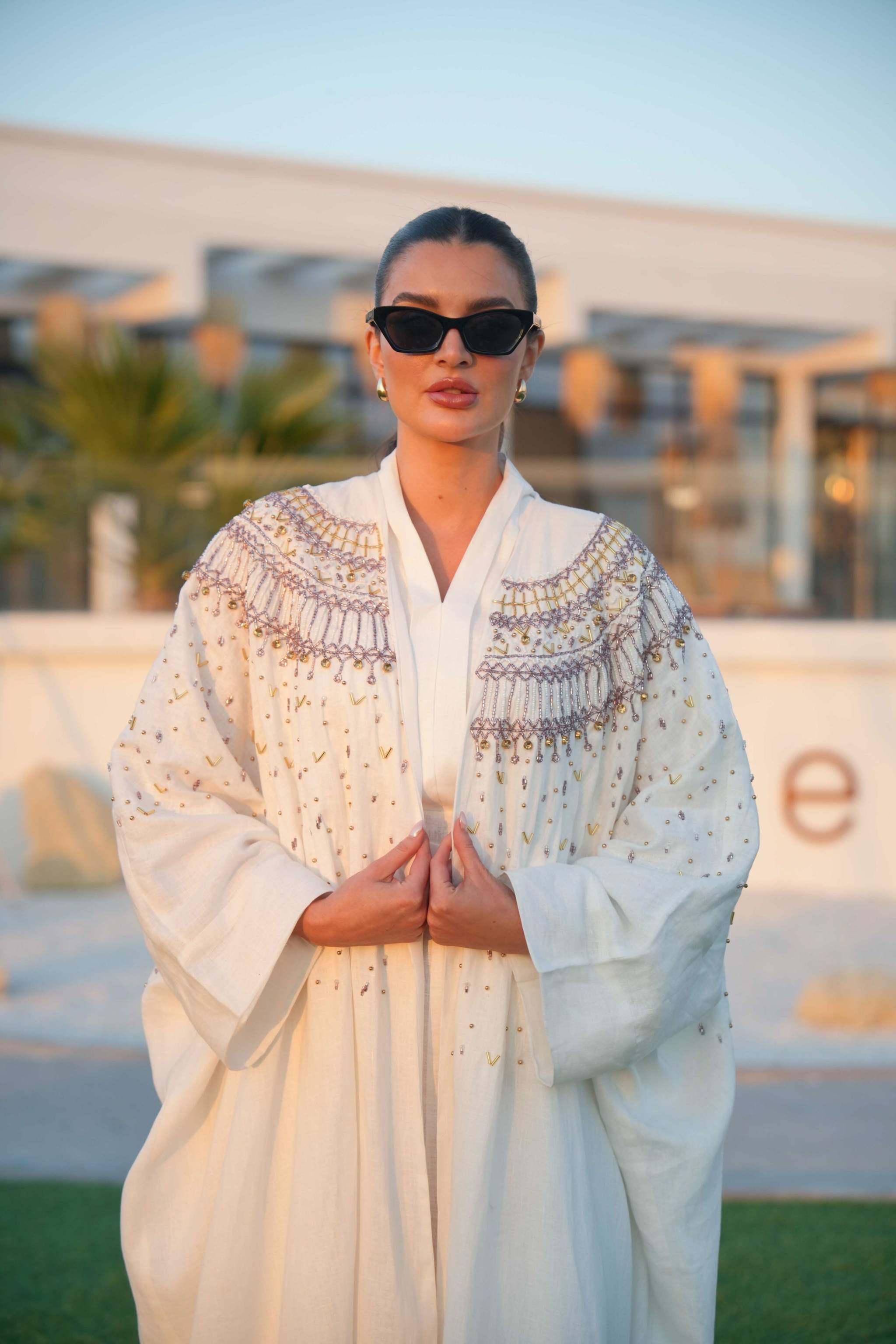 Elegant white Aya abaya with intricate gold and soft pink embroidery on shoulders and sleeves, worn by woman with sunglasses outdoors