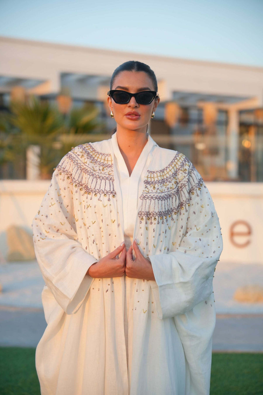 Elegant white Aya abaya with intricate gold and soft pink embroidery on shoulders and sleeves, worn by woman with sunglasses outdoors