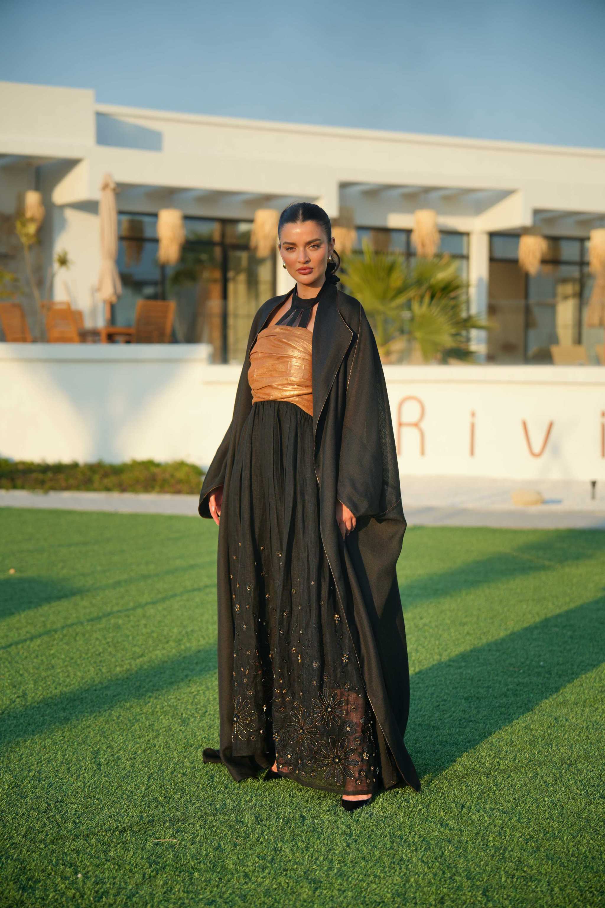 Woman wearing Golden Touch ensemble featuring black bisht with golden embroidery and black to gold gradient linen jumpsuit outdoors on grass