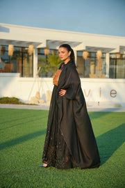 Woman wearing a black khesha bisht with golden touch embroidery paired with a black to golden linen jumpsuit featuring subtle golden details, styled outdoors.