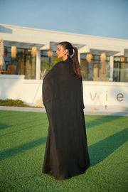 Woman wearing a black khesha bisht with golden touch embroidery paired with a black to golden linen jumpsuit featuring subtle golden details, styled outdoors.