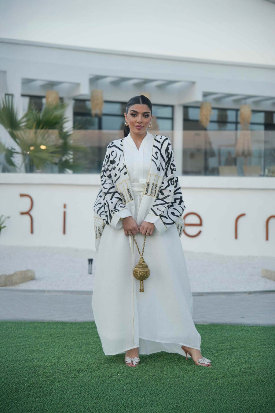 woman wearing elegant white bisht with bold black Arabic calligraphy embroidery on sleeves and upper bodice standing outdoors