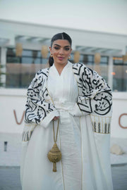 woman wearing elegant white bisht with bold black Arabic calligraphy embroidery on sleeves and upper bodice standing outdoors