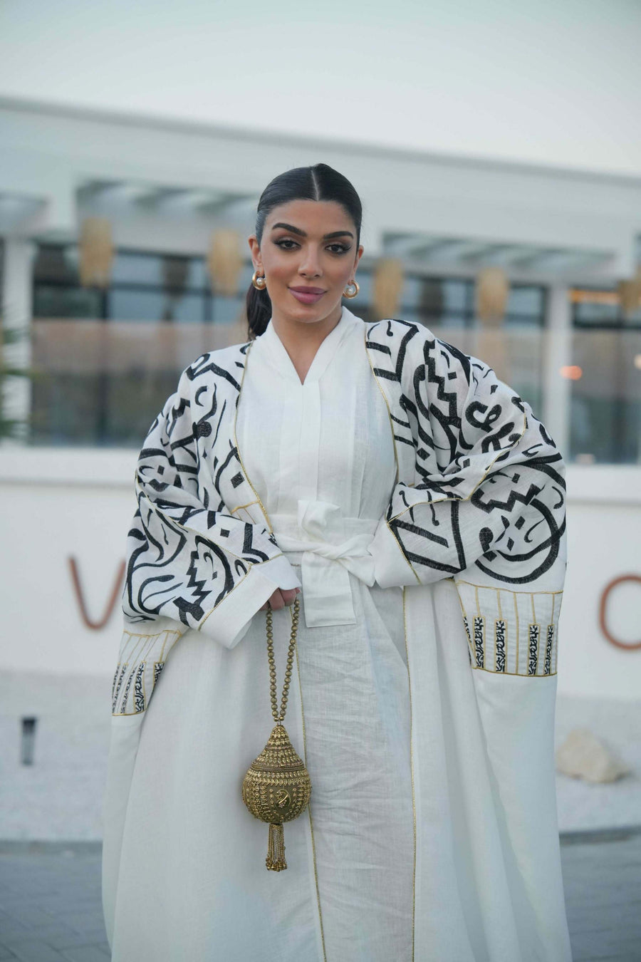 woman wearing elegant white bisht with bold black Arabic calligraphy embroidery on sleeves and upper bodice standing outdoors