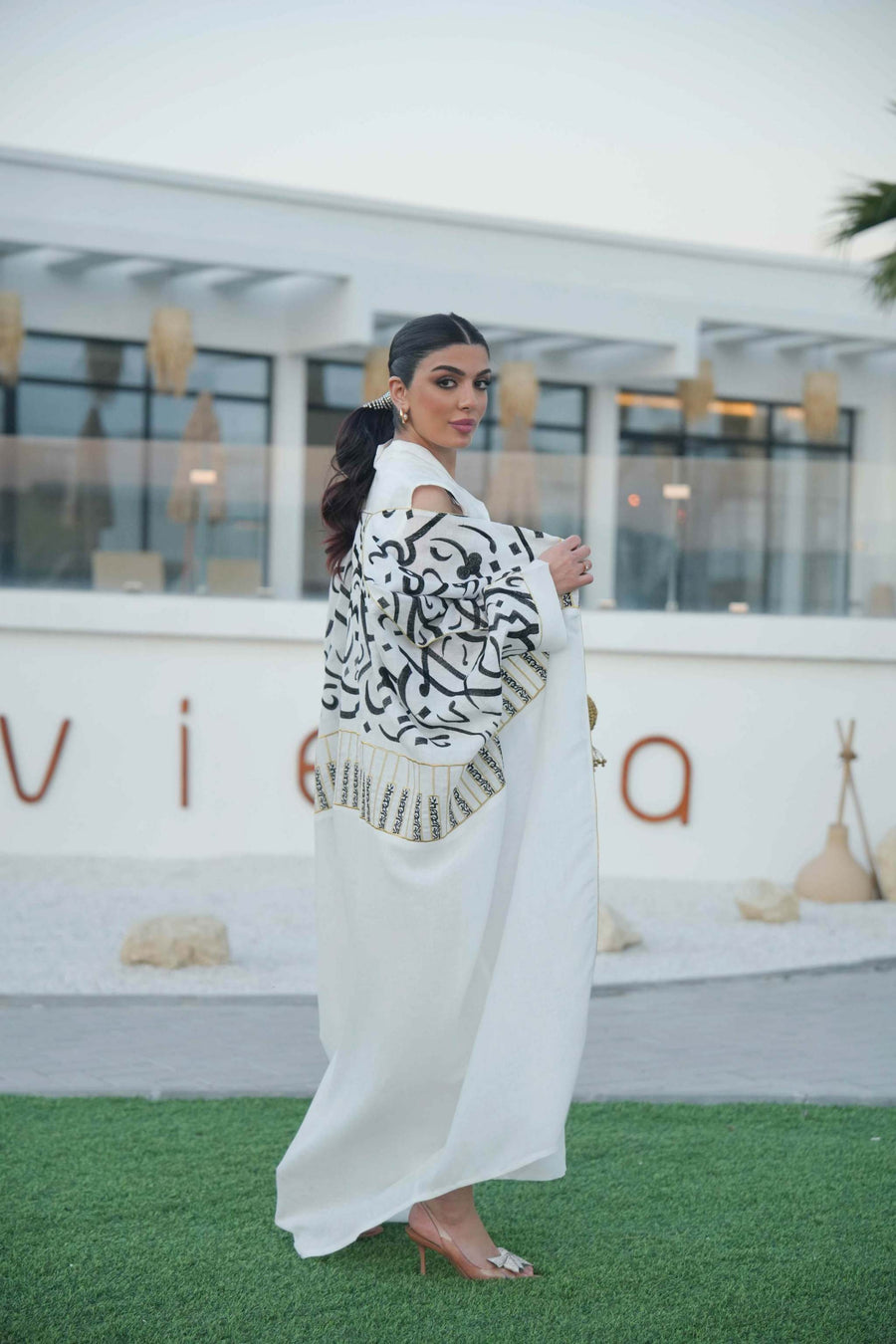 woman wearing elegant white bisht with bold black Arabic calligraphy embroidery on sleeves and upper bodice standing outdoors