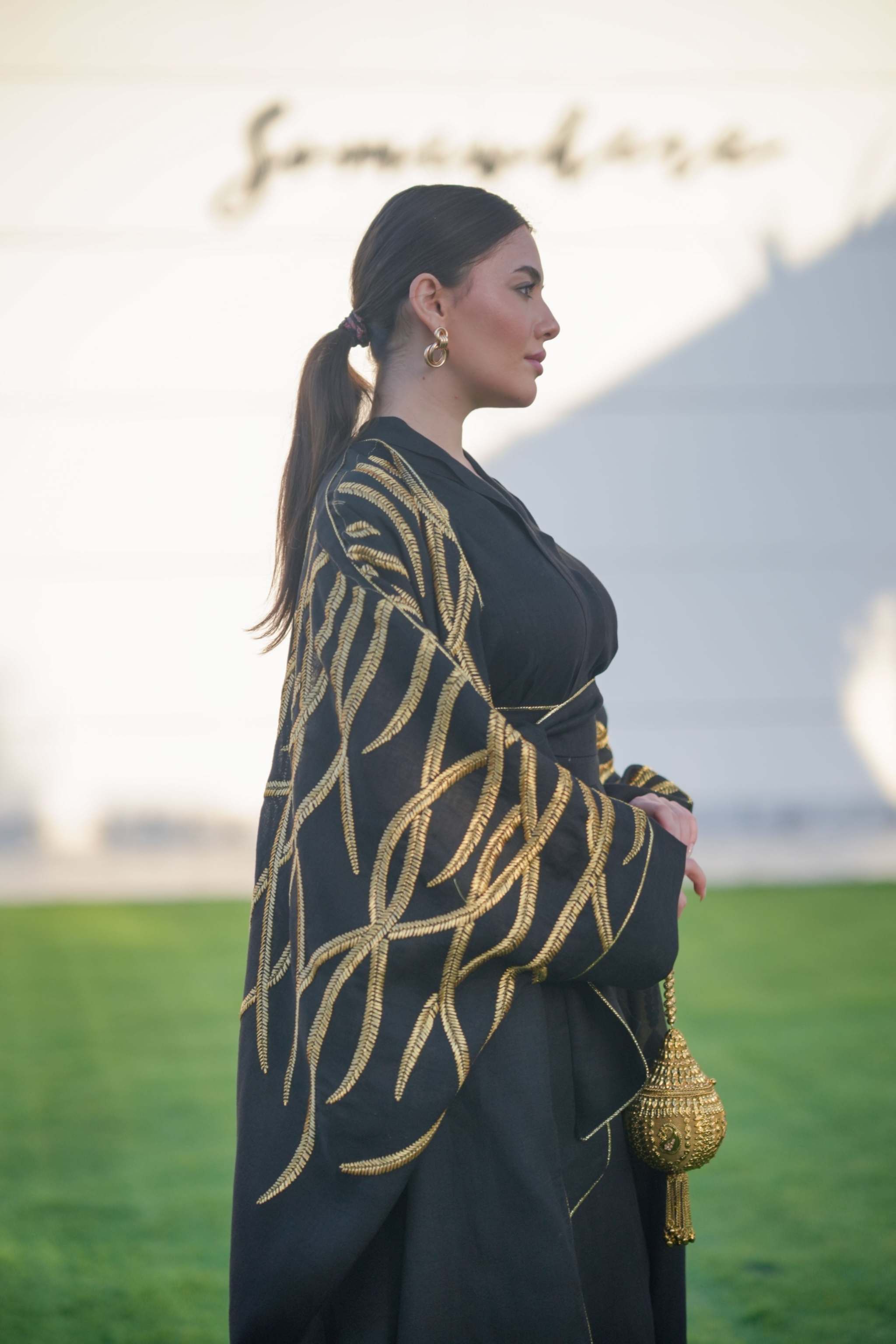 Woman wearing black linen Bisht with bold gold embroidery in leaf-like pattern, posing outdoors with a gold tassel handbag.