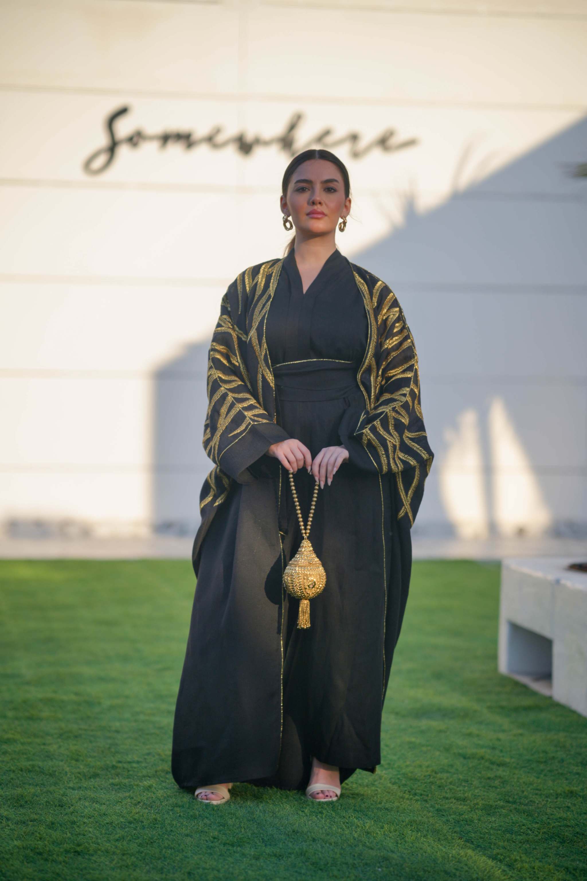 Woman wearing a black linen bisht with bold gold leaf embroidery and wide sleeves, holding a gold beaded bag, standing outdoors on grass.