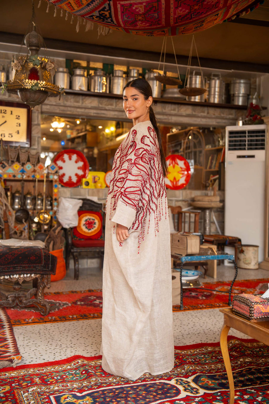 Woman wearing beige bisht with bold red embroidery on shoulders and sleeves called Crimson Bloom in cozy indoor setting