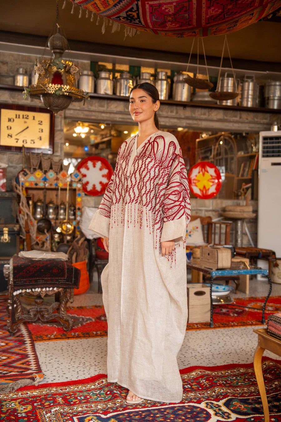 Woman wearing beige bisht with bold red embroidery on shoulders and sleeves called Crimson Bloom in cozy indoor setting