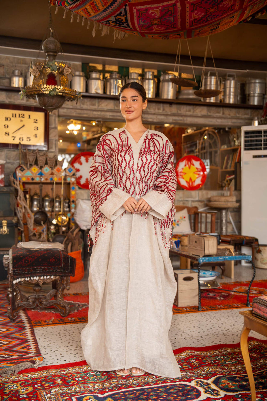 Woman wearing beige bisht with bold red embroidery on shoulders and sleeves called Crimson Bloom in cozy indoor setting