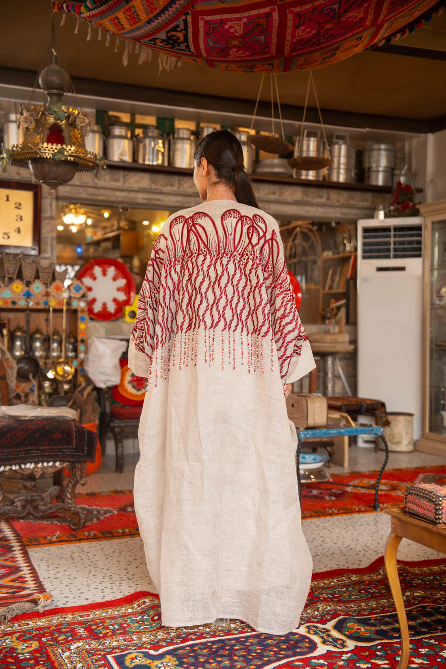 Woman wearing beige bisht with bold red embroidery on shoulders and sleeves called Crimson Bloom in cozy indoor setting