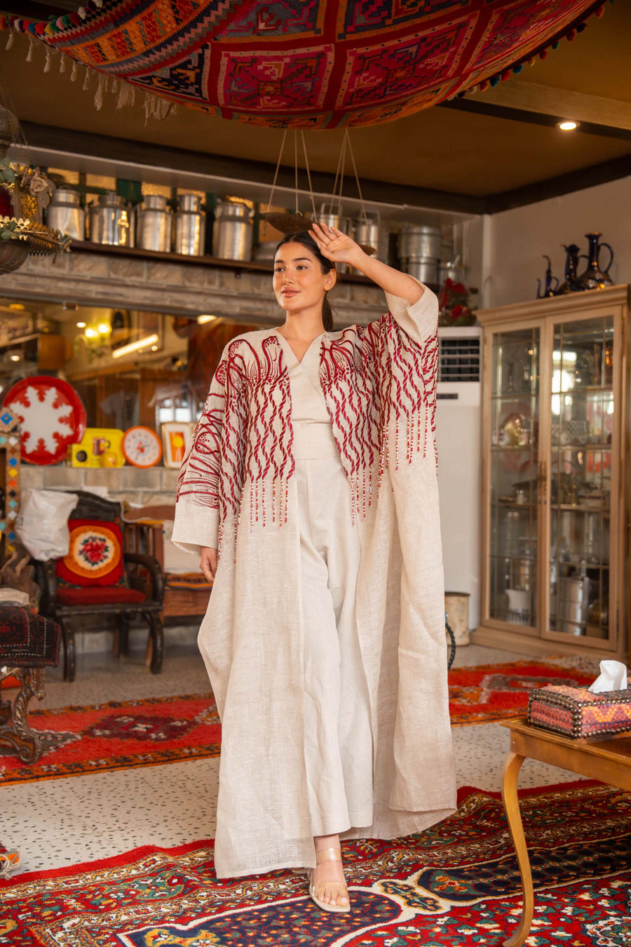 Woman wearing beige bisht with bold red embroidery on shoulders and sleeves called Crimson Bloom in cozy indoor setting