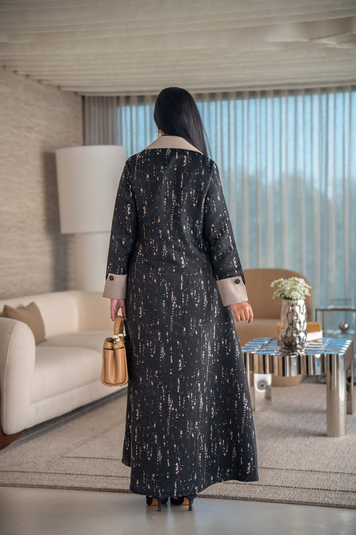 woman wearing elegant black and beige patterned Ashira abaya holding tan handbag in modern living room
