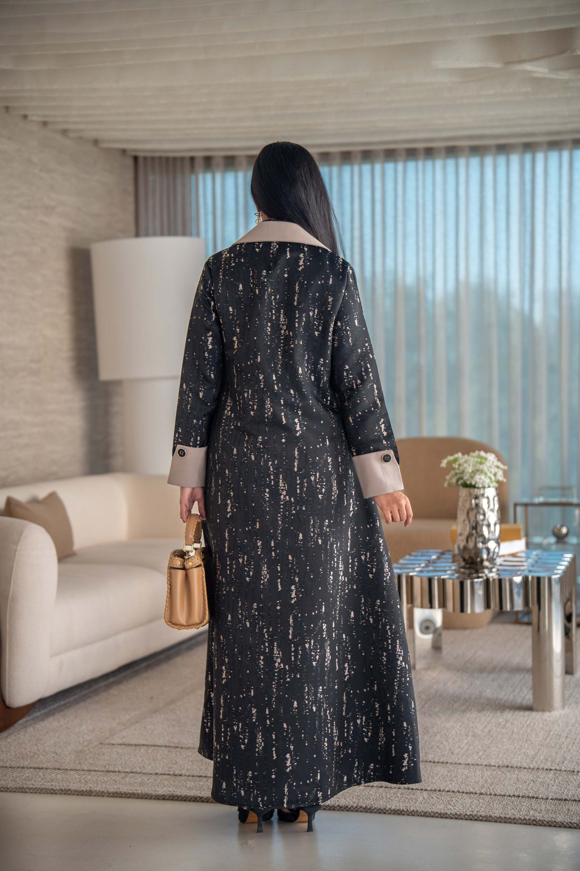 woman wearing elegant black and beige patterned Ashira abaya holding tan handbag in modern living room
